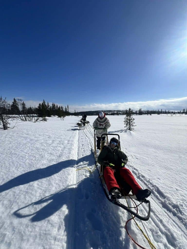 09349d50-5698-4771-91ba-06b0369b63ef (1) To personer sitter på en hundeslede som kjører gjennom et snødekt landskap under en klarblå himmel, med trær spredt i det fjerne.