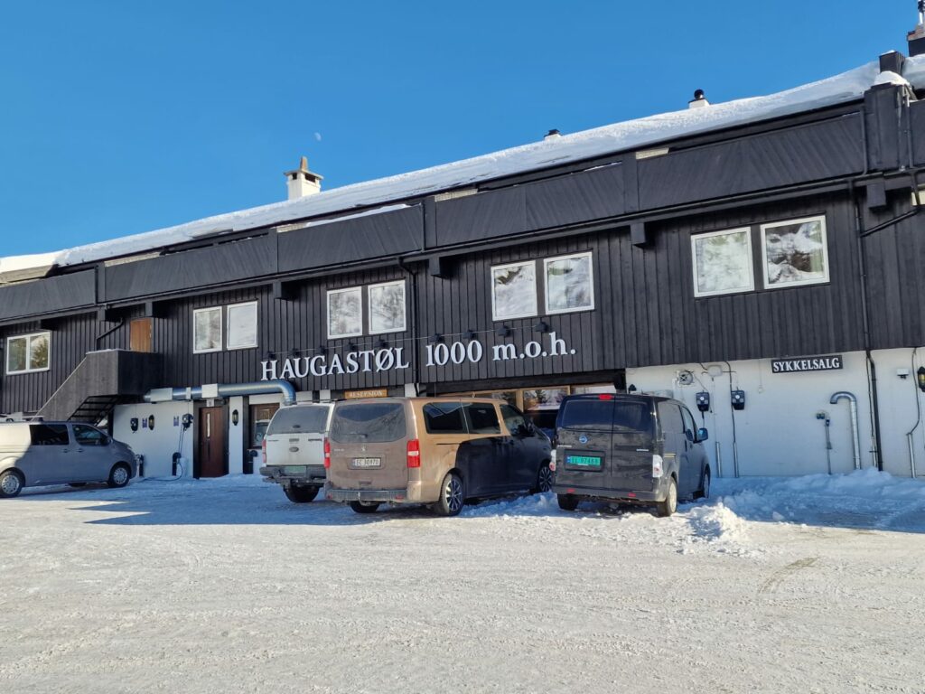 En svart bygning med skiltet "Haugastøl 1000 m.o.h." har snødekt mark og flere parkerte biler foran.