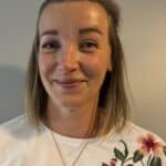 En kvinne med skulderlangt blondt hår, kledd i en hvit blomstret skjorte og et halskjede, smiler foran en ensfarget grå bakgrunn.