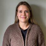 En kvinne med langt hår smiler til kameraet. Hun har på seg en brun, strukturert cardigan over en svart skjorte og står foran en ensfarget, lys vegg.