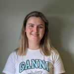 En person med skulderlangt blondt hår smiler til kameraet. Vedkommende har på seg en hvit T-skjorte med påskriften «GANNI» og et haimotiv, og står foran en ensfarget, lys vegg.
