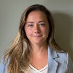 En kvinne med langt, lysebrunt hår, kledd i en lyseblå blazer og en kremfarget topp, står foran en ensfarget, lys vegg og ser inn i kameraet med et nøytralt uttrykk.