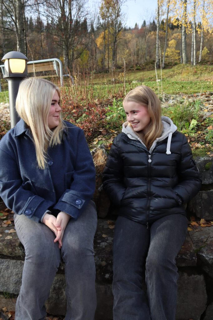 20240919_101230050_iOS-scaled To mennesker sitter på en steinmur utendørs om høsten, vendt mot hverandre og smilende, med trær og løvfall i bakgrunnen.