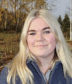 En kvinne med langt blondt hår og en blå frakk smiler ute i en park med høsttrær og en statue i bakgrunnen.
