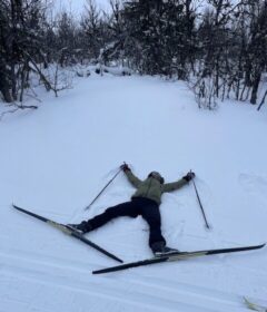 En person i vinterklær ligger på ryggen i snøen med ski og staver spredt ut, omgitt av trær.