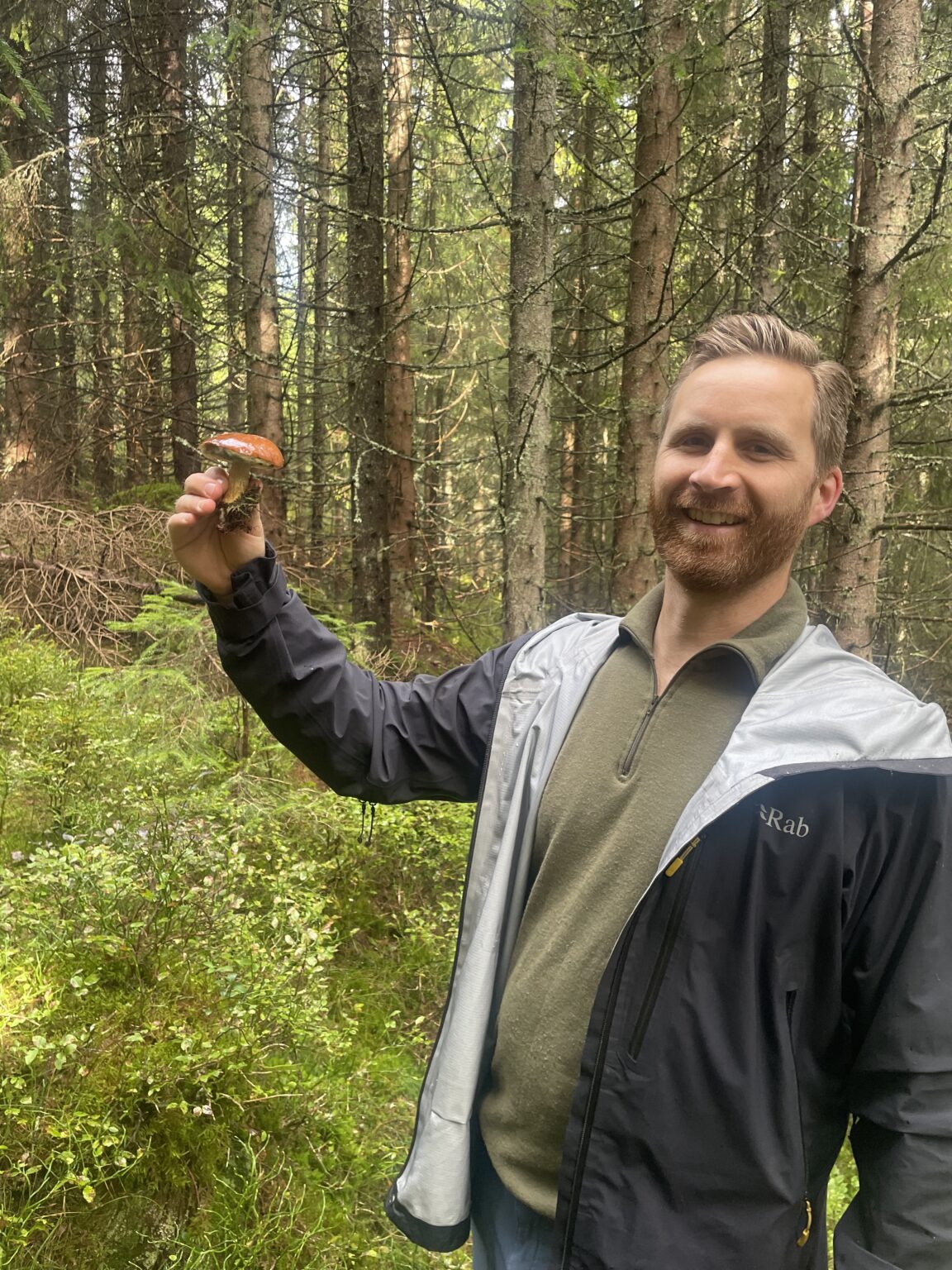 En mann står i en skog med en sopp i høyre hånd og smiler til kameraet.