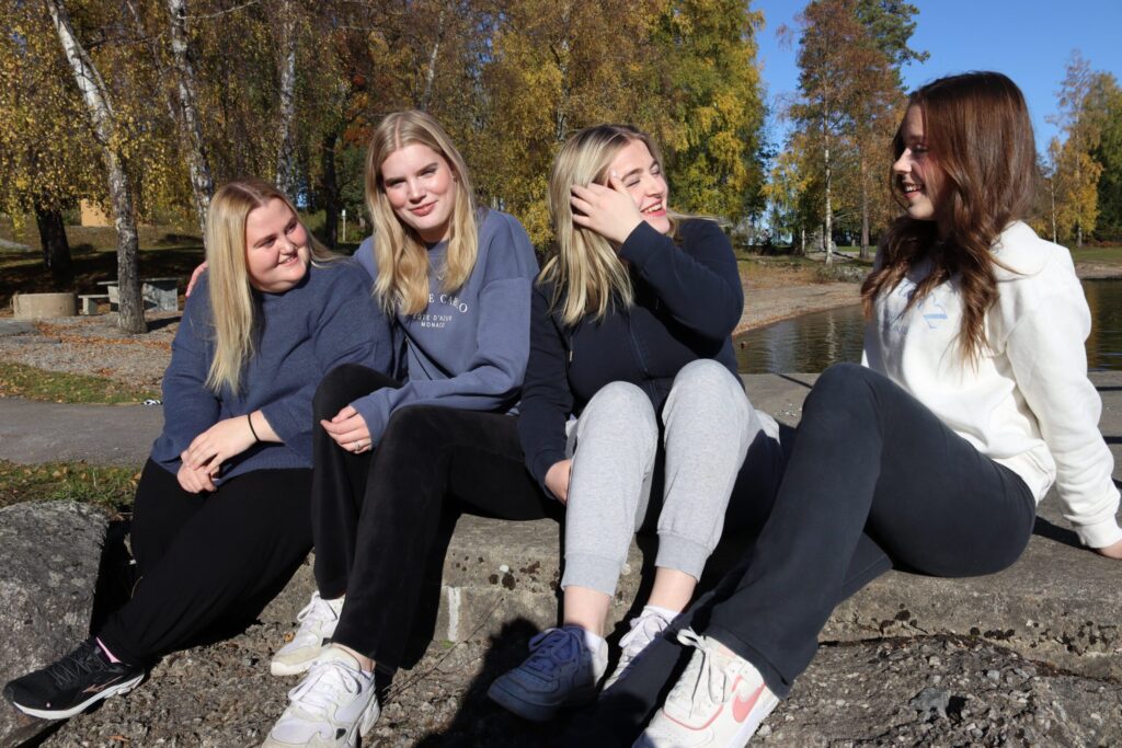 Fire unge kvinner sitter på en lav steinmur utendørs om høsten, lettkledde og smilende mens de snakker sammen. I bakgrunnen ser vi trær med gule blader og et vann.
