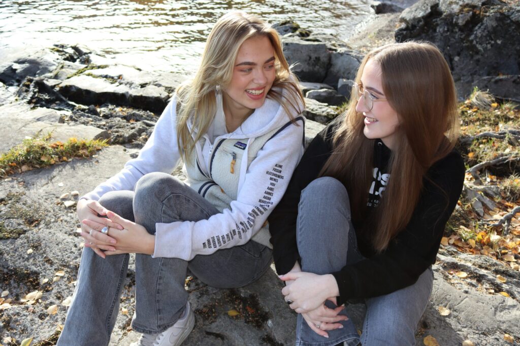 To unge kvinner sitter på steiner ved vannet, snakker og smiler til hverandre. Begge er lettkledde i jeans og hettegensere.