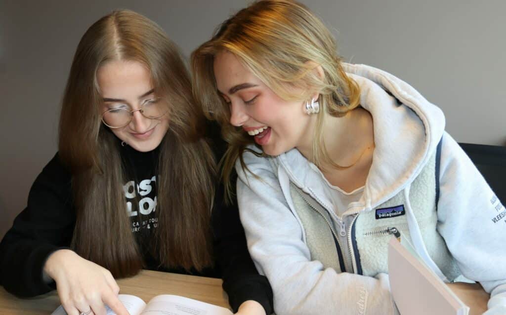 To unge kvinner sitter ved et bord og ser på en oppslått bok sammen, den ene smiler og den andre peker på sidene.