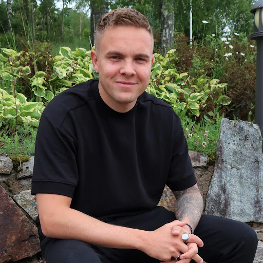 heine En person med kort hår og svart t-skjorte sitter utendørs foran en steinmur og grønne planter og smiler til kameraet.