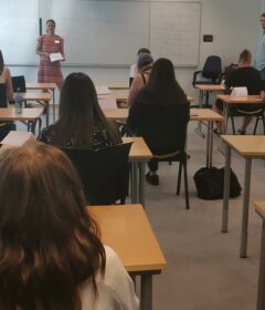 Elevene sitter ved pulter i et klasserom vendt mot en tavle, mens en kvinne står foran og snakker, og en mann står like ved.