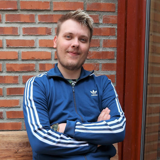 robin-marco En person med kort, blondt hår og lyst ansiktshår sitter på en trebenk foran en murvegg, iført blå Adidas-jakke og armene i kors.