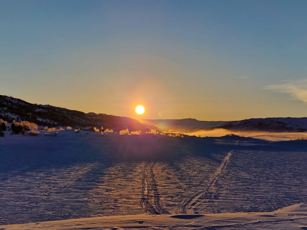 Solen går ned over et snødekt landskap med åser i bakgrunnen og ski- eller skuterspor i snøen.