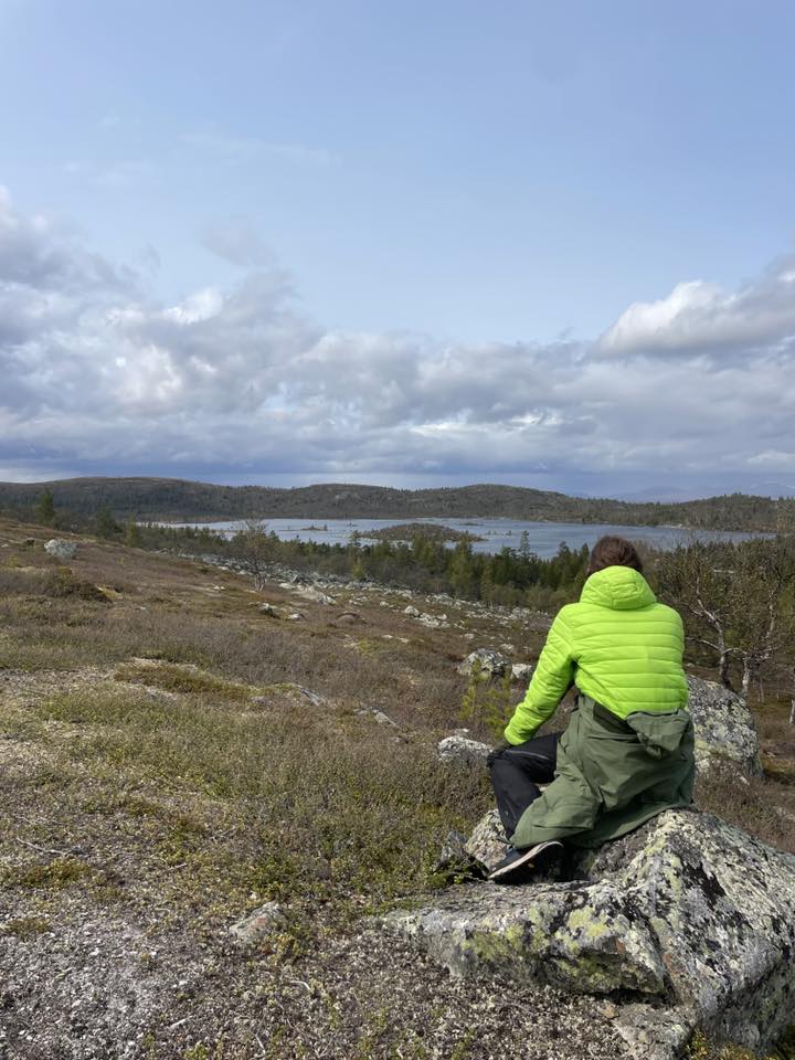 En person i lysegrønn jakke sitter på en stein, vendt mot et naturskjønt landskap med en innsjø, åser, spredte trær og en skyet himmel.