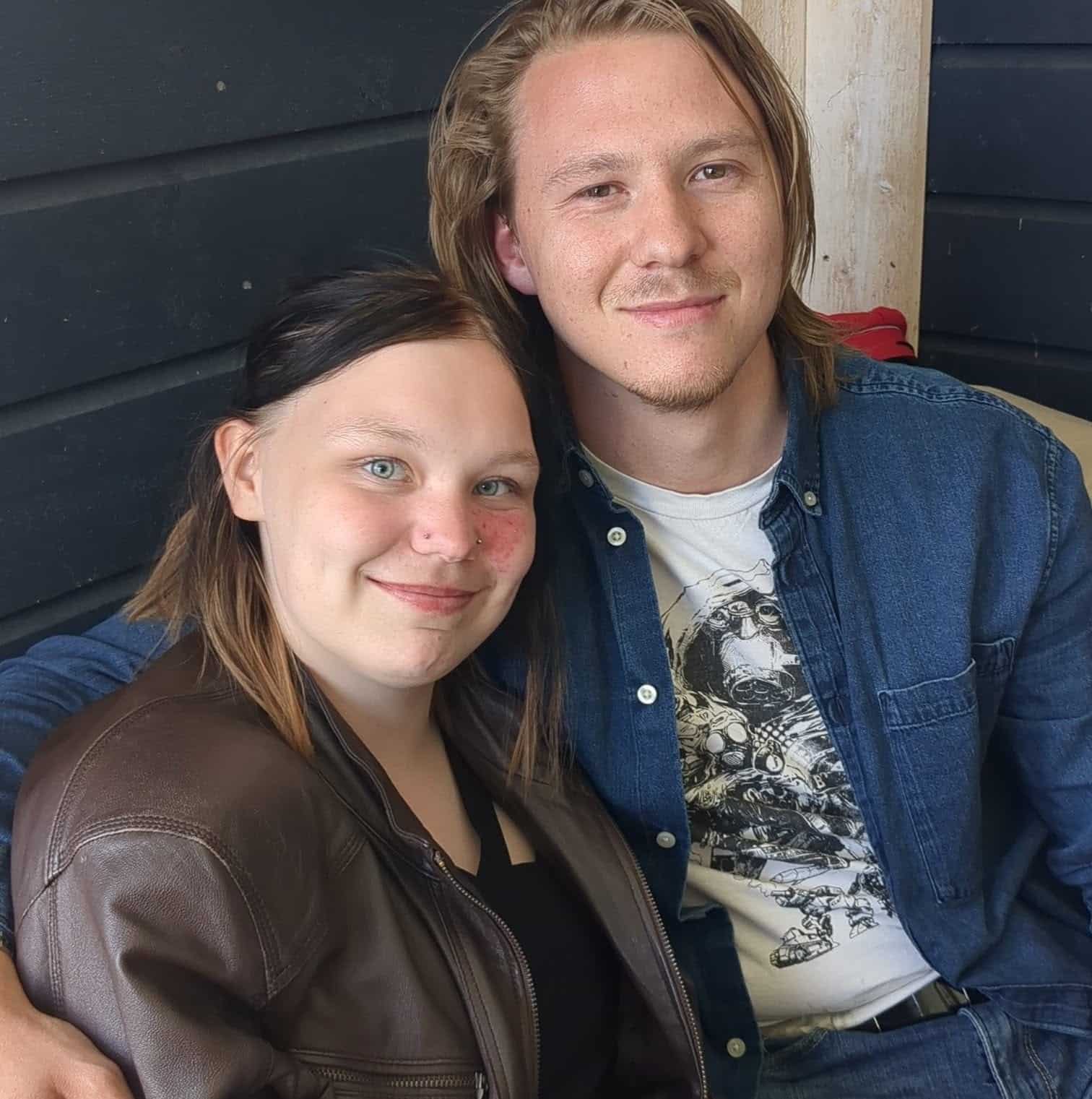 En kvinne og en mann sitter tett sammen, begge smiler mot kameraet. Kvinnen har på seg en brun jakke, og mannen har på seg en blå skjorte over en t-skjorte med trykk.