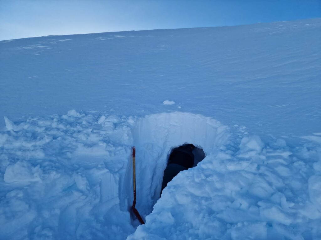 En snøhuleinngang gravd inn i en snødekt skråning med en spade synlig i nærheten og en person delvis inne i tunnelen.