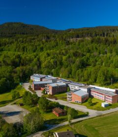 Flyfoto av en moderne skolecampus med flere mursteinsbygninger omgitt av grønne trær og åkrer, mot en skogkledd åsside under en klar blå himmel.