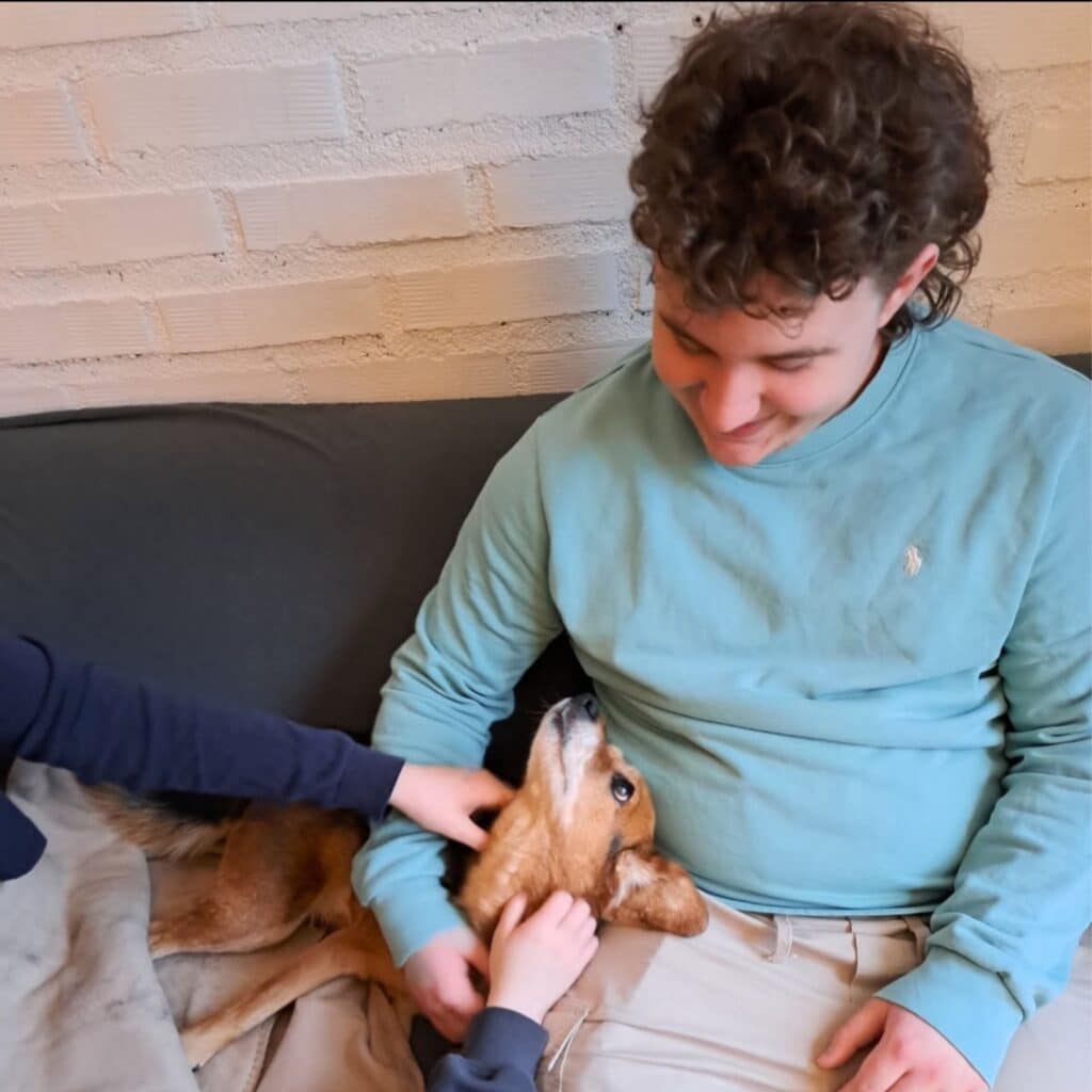 Design uten navn En person i lyseblå skjorte sitter i en sofa og smiler ned på en hund som ligger ved siden av, mens en annen person klapper hunden.