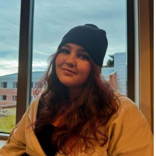 Design uten navn (3) En person med langt, brunt hår, svart lue og lys jakke sitter innendørs foran et vindu med bygninger og en overskyet himmel utenfor.