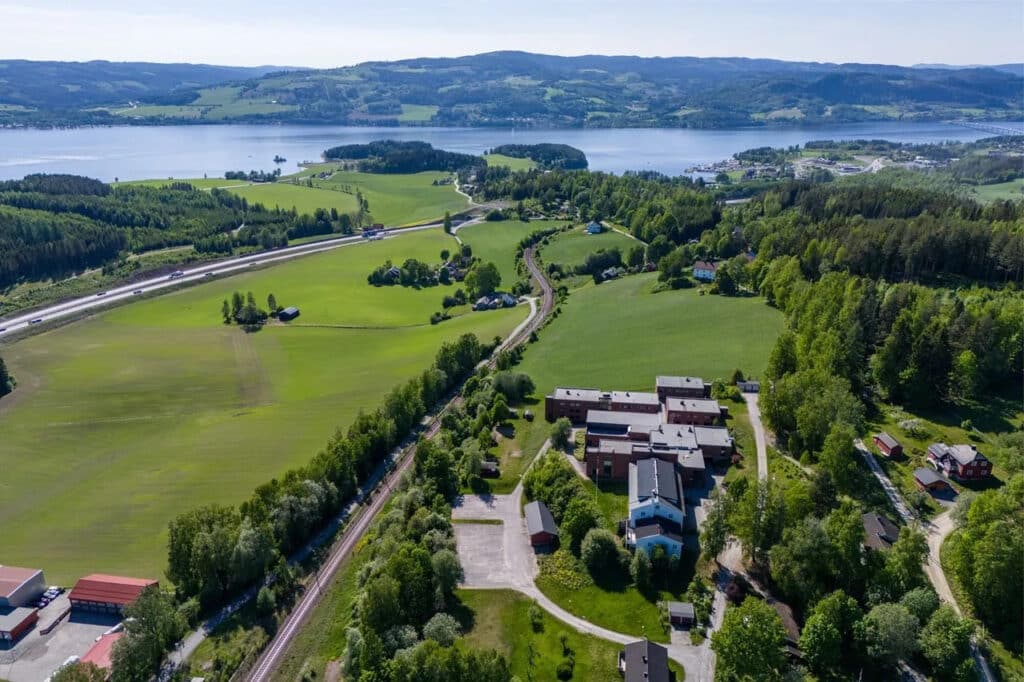 Flyfoto av et ruralt landskap med åkrer, spredte bygninger, en jernbane og en innsjø omgitt av åser i bakgrunnen under en klar himmel.