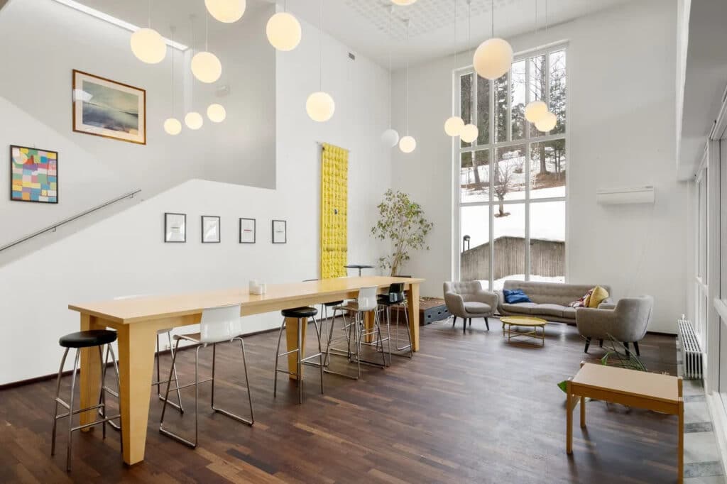 Moderne, romslig stue med stor takhøyde, store vinduer, hengende globuser, et langt spisebord med krakker og en sittegruppe med lenestoler og en sofa.