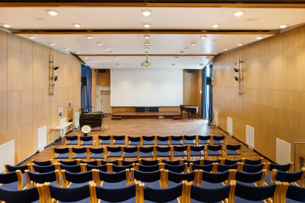 IMG_5878 Rader av tomme, blå stoler står foran en scene med et stort, hvitt lerret, et piano og trepanel på veggene i et godt opplyst auditorium.