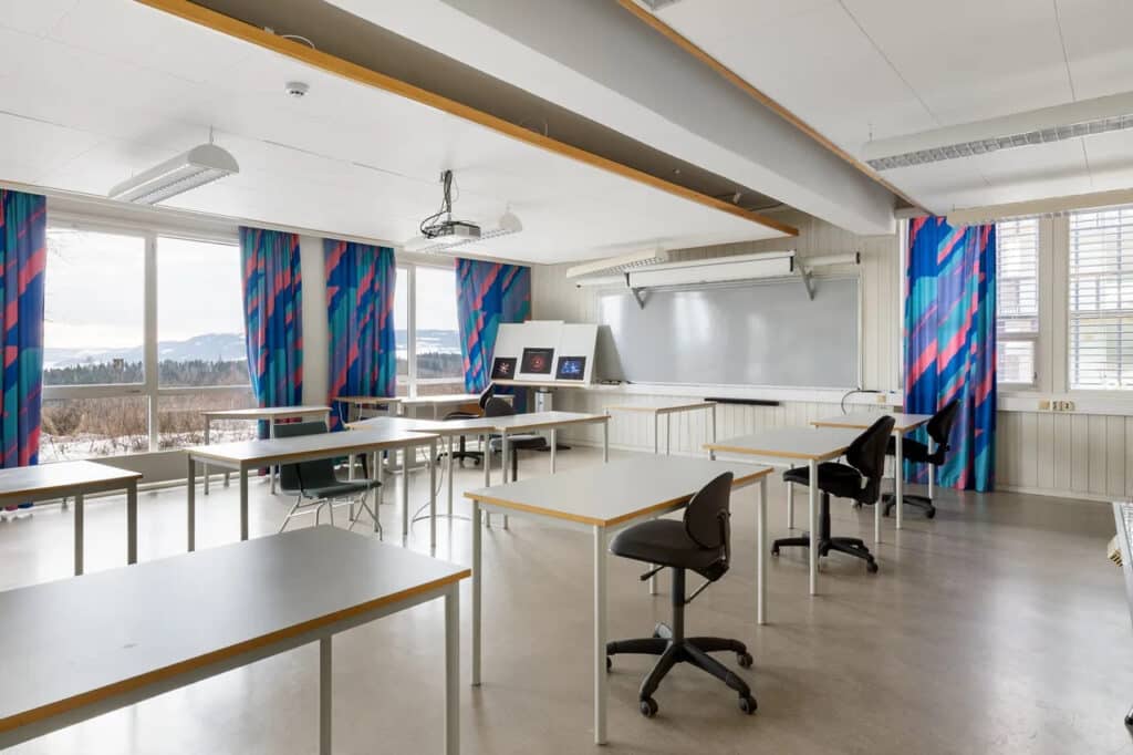 Tomt klasserom med flere bord og stoler, store vinduer med blå og rosa gardiner, whiteboardtavle og en projektor i taket.