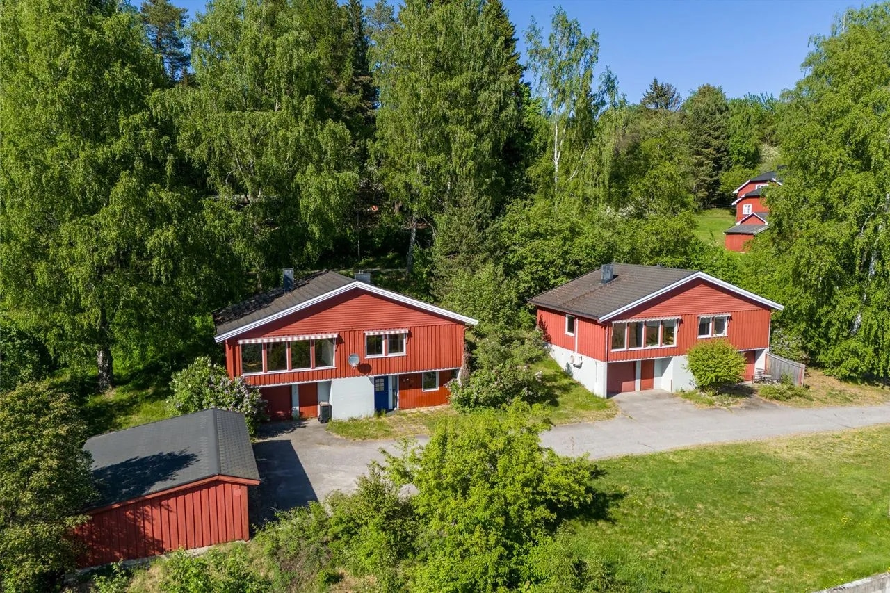 Flyfoto av to røde hus med hvite kanter, et lite rødt skur og grønne trær og gress rundt, under en klar blå himmel.