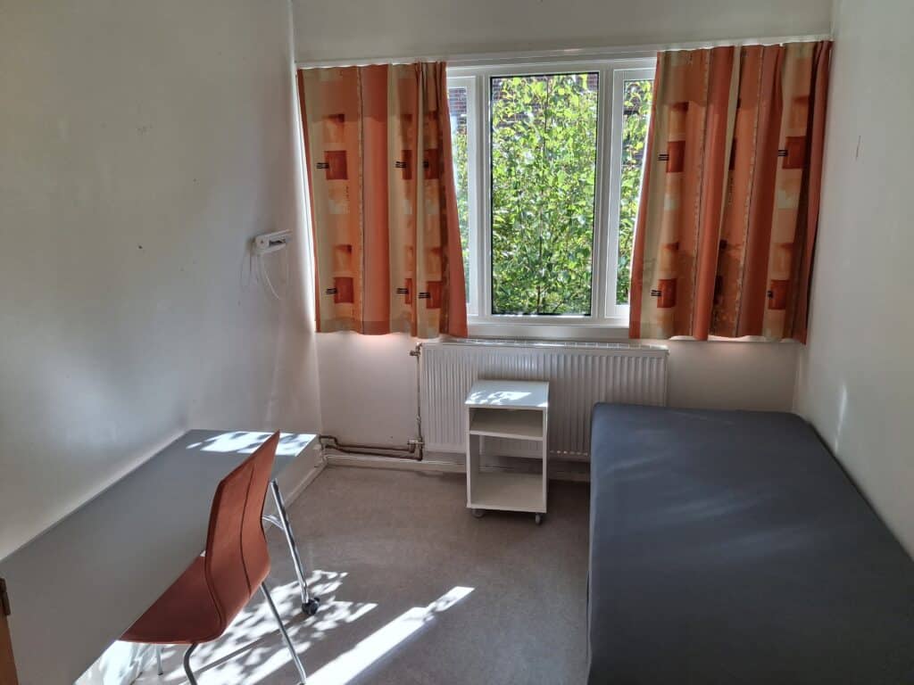 Et lite, enkelt rom med en seng, et skrivebord, en stol og et vindu med oransje gardiner. Sollyset strømmer inn og lyser opp møblene og det beige teppet.