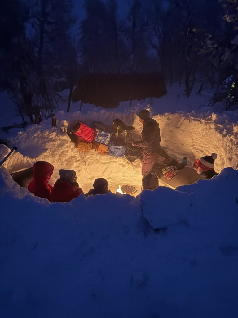 a7aadda5-3ae8-401b-97c3-1954f7d2467e En gruppe mennesker sitter rundt et bål i en snødekt grop om natten, omgitt av snøbanker og vinterutstyr, med trær og et gapahuk i bakgrunnen.