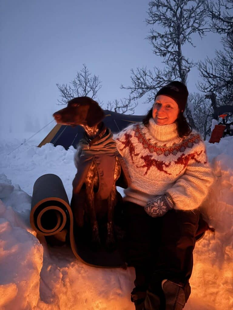 b17864d4-17d6-4fe0-a8ac-ee27c18122a8 En kvinne i strikkegenser sitter ved siden av en hund på en yogamatte ved et bål i et snødekt utemiljø, med nakne trær og et telt i bakgrunnen.