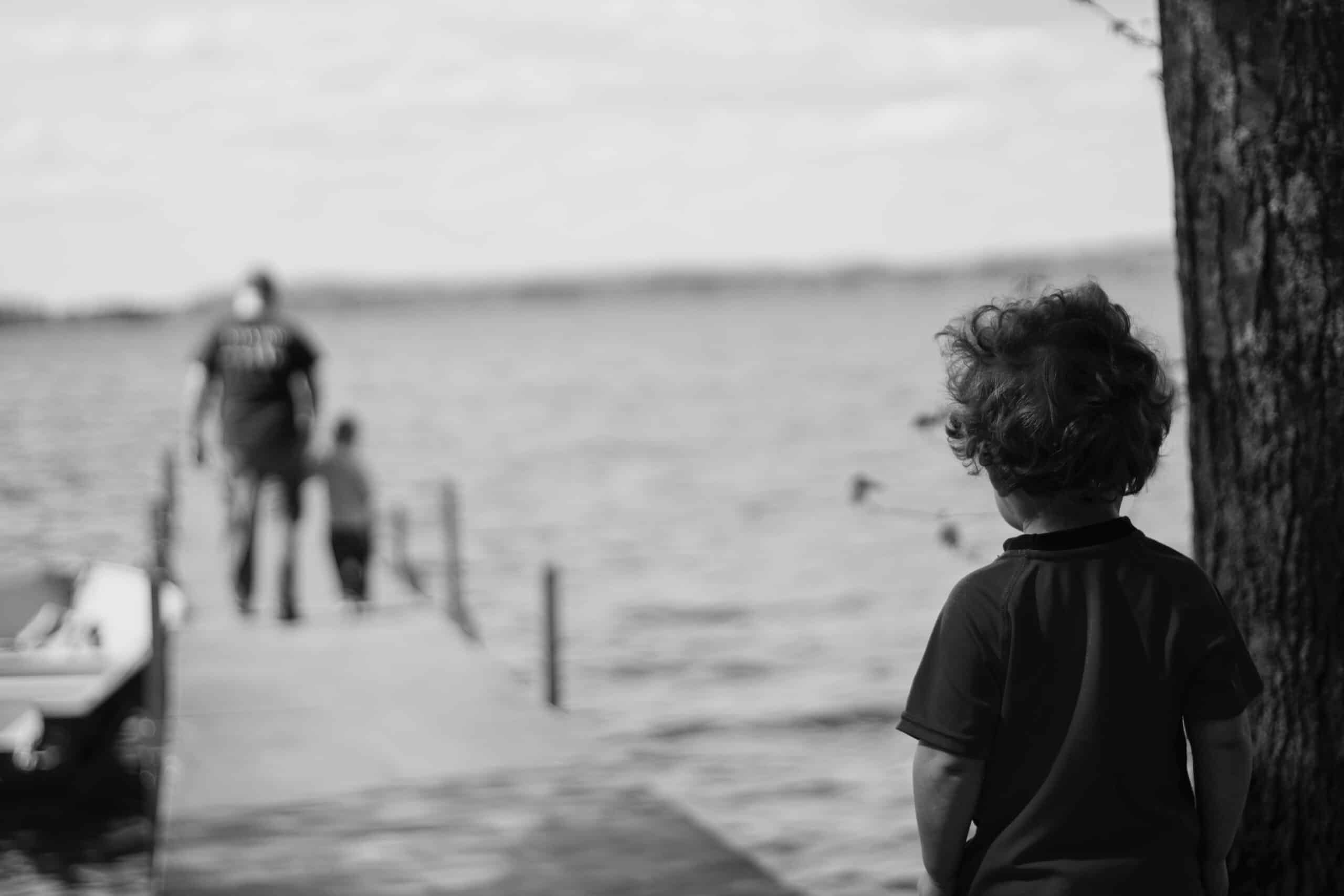 Et barn står ved et tre og ser på to personer som går på en brygge som strekker seg ut i en innsjø; bildet er i svart-hvitt.
