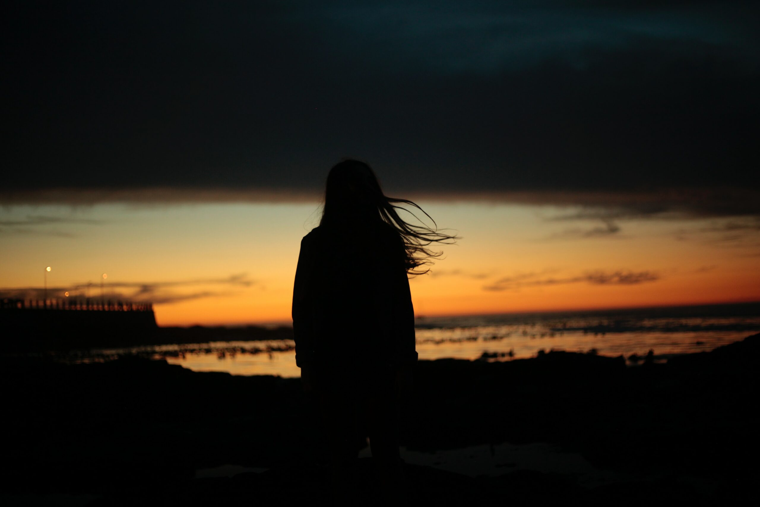 Silhuett av en person som står i fjæra ved solnedgang, med rolig vann og en brygge i bakgrunnen.